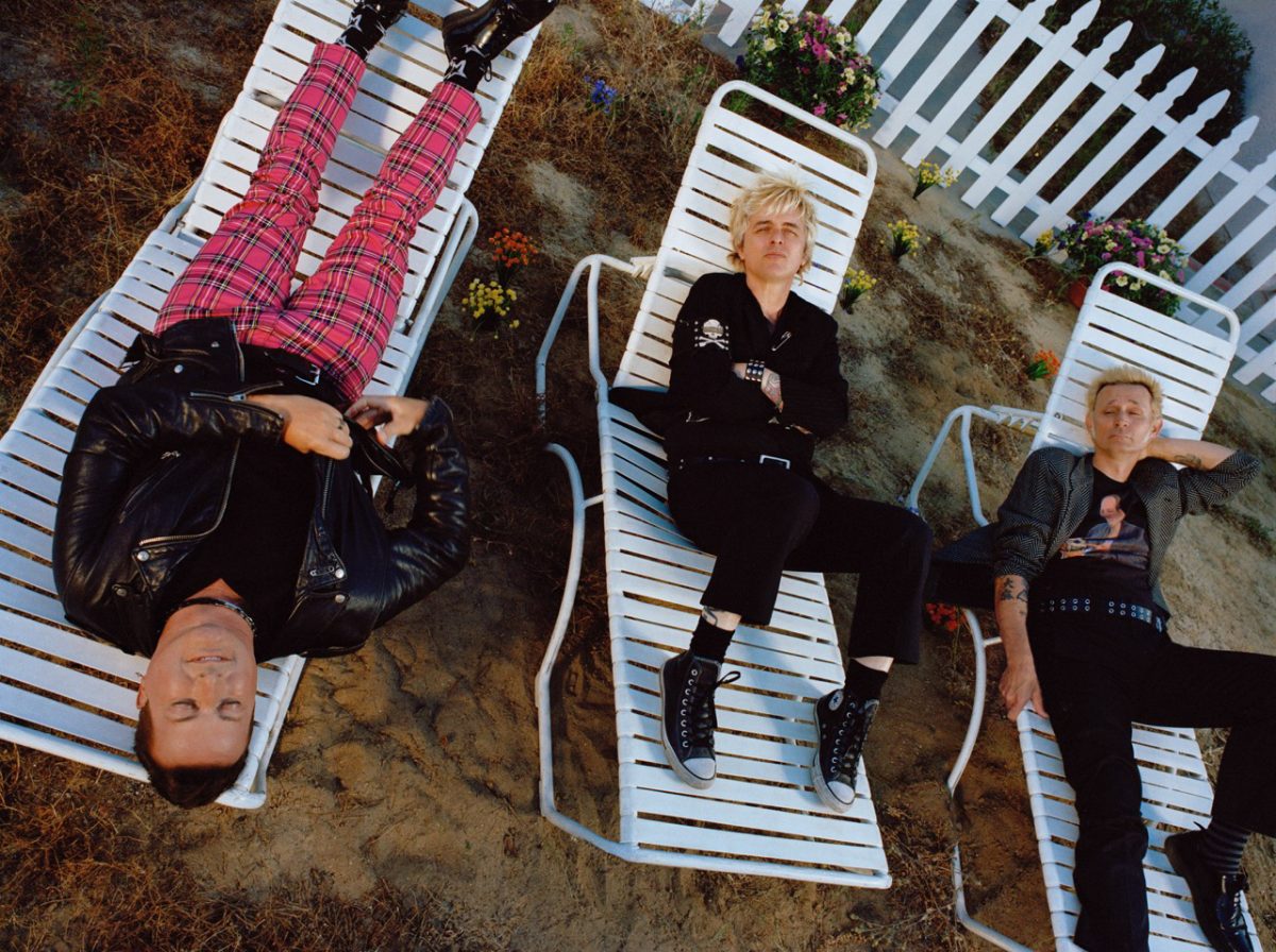 Members of Green Day lying on their backs on white lawn chairs. One member is lying upside down while the other two are upright with their legs bent. They are located in a garden with dead grass, sand, patchy flowers, and a white picket fence.