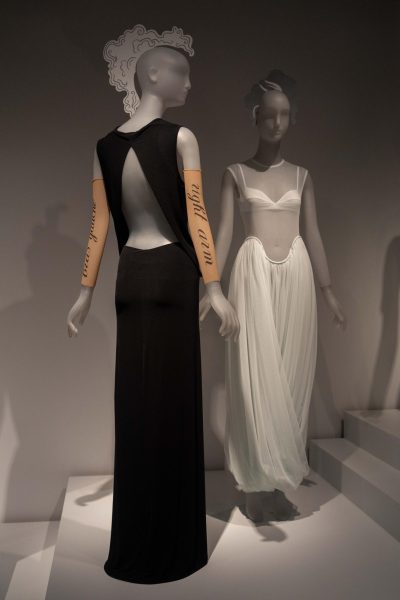 Two mannequins on display. One wears wears a black dress with a back cutout, nude arm warmers and a paper headpiece. The other wears a white long-sleeve dress with a sheer top and a white brassiere underneath.