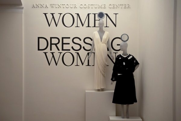 Two mannequins in front of a wall that says “ANNA WINTOUR COSTUME CENTER” and “WOMEN DRESSING WOMEN.” One mannequin wears a white dress and one wears a black dress.