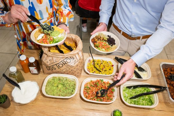 Two people, each holding a bowl and a spoon in their hands, take food from a table filled with sauces and food including corn, tomatos, guacomole, lettuce and cheese.