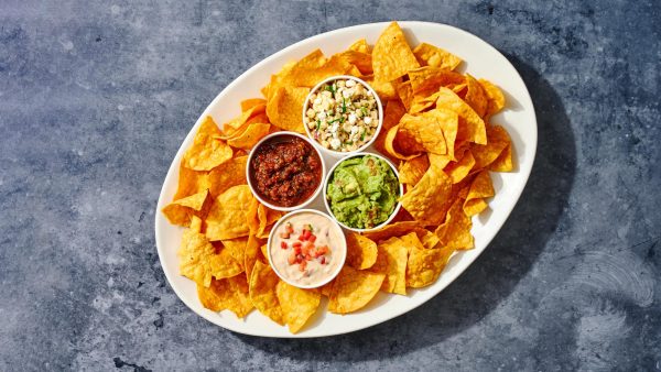 A plate filled with tortilla chips and four types of sauces at the middle is placed on a brightly lit blue table.