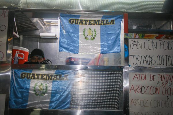 A food cart with the blue-and-white Guatemalan flag. A person is peeking through the window of the cart and there are paper menus attached to the front of the cart.