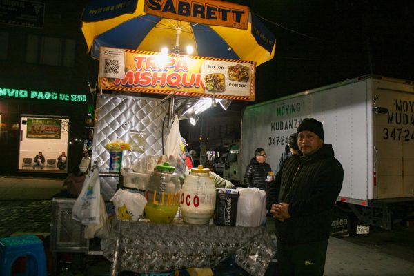 A food cart with a blue and yellow umbrella on the top. It has a board that says "TRIPA MISHQUI" and has pictures of food. The owner is standing next to the cart.