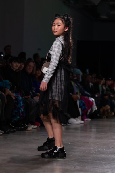 A model walking down a runway with a sheer black dress over a gray plaid dress. The model also wears a fluffy cat-ear headband and carries a small porcupine cross-body bag.