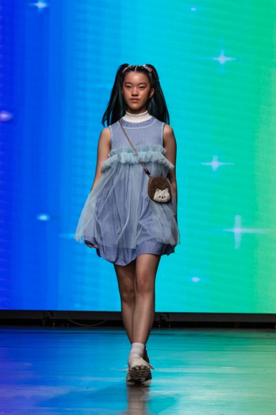 A model walking down a runway with a blue dress with a white turtleneck underneath and a small porcupine cross-body bag.