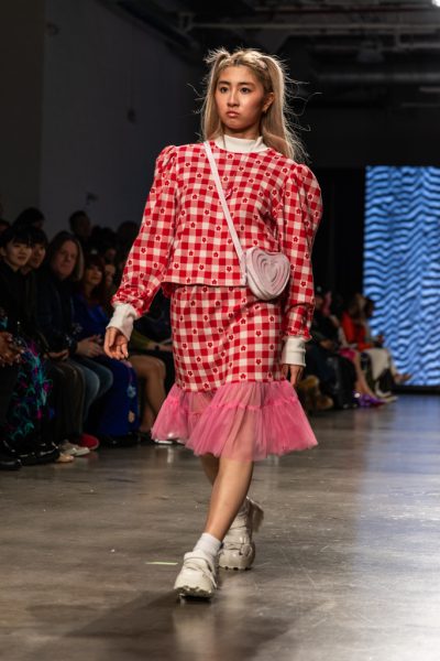 A model walking down a runway with a red-and-white plaid shirt, a skirt with a pink tulle trim and a light pink cross-body bag.
