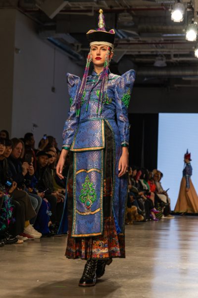 A model walking down a runway with a traditional blue Mongolian-inspired top and headpiece.
