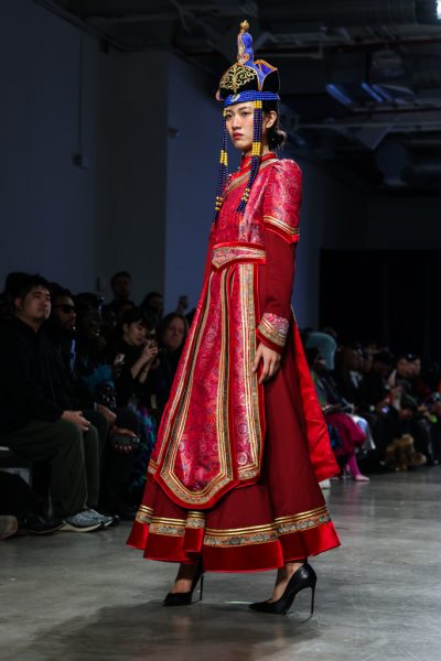 A model walking down a runway with a red dress and a hat with beaded strands and gold detailing.