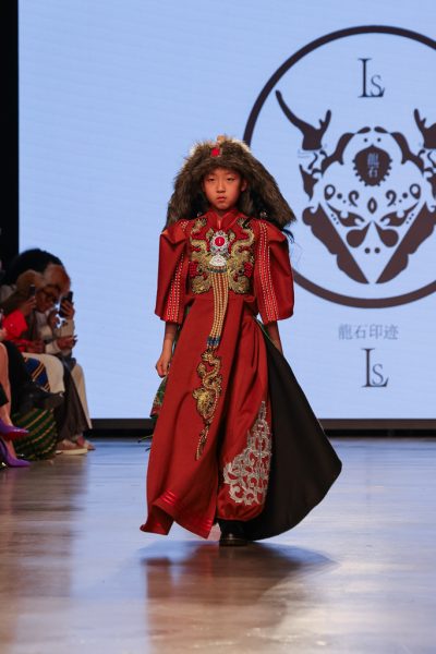 A model walking down a runway with a Mongolian-inspired fur headpiece and red dress with golden embroidery.