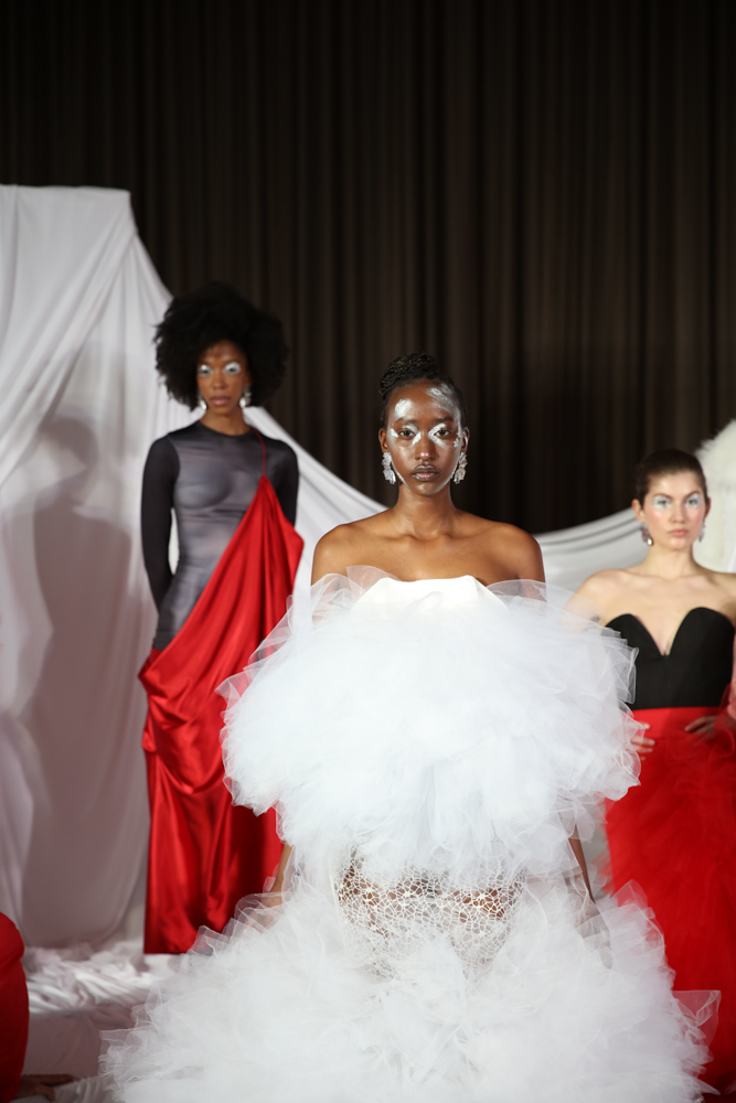 Three models stand in front of a white backdrop. The center model wears a white dress with poofy tulle, and the two models behind her wear black-and-red dresses.