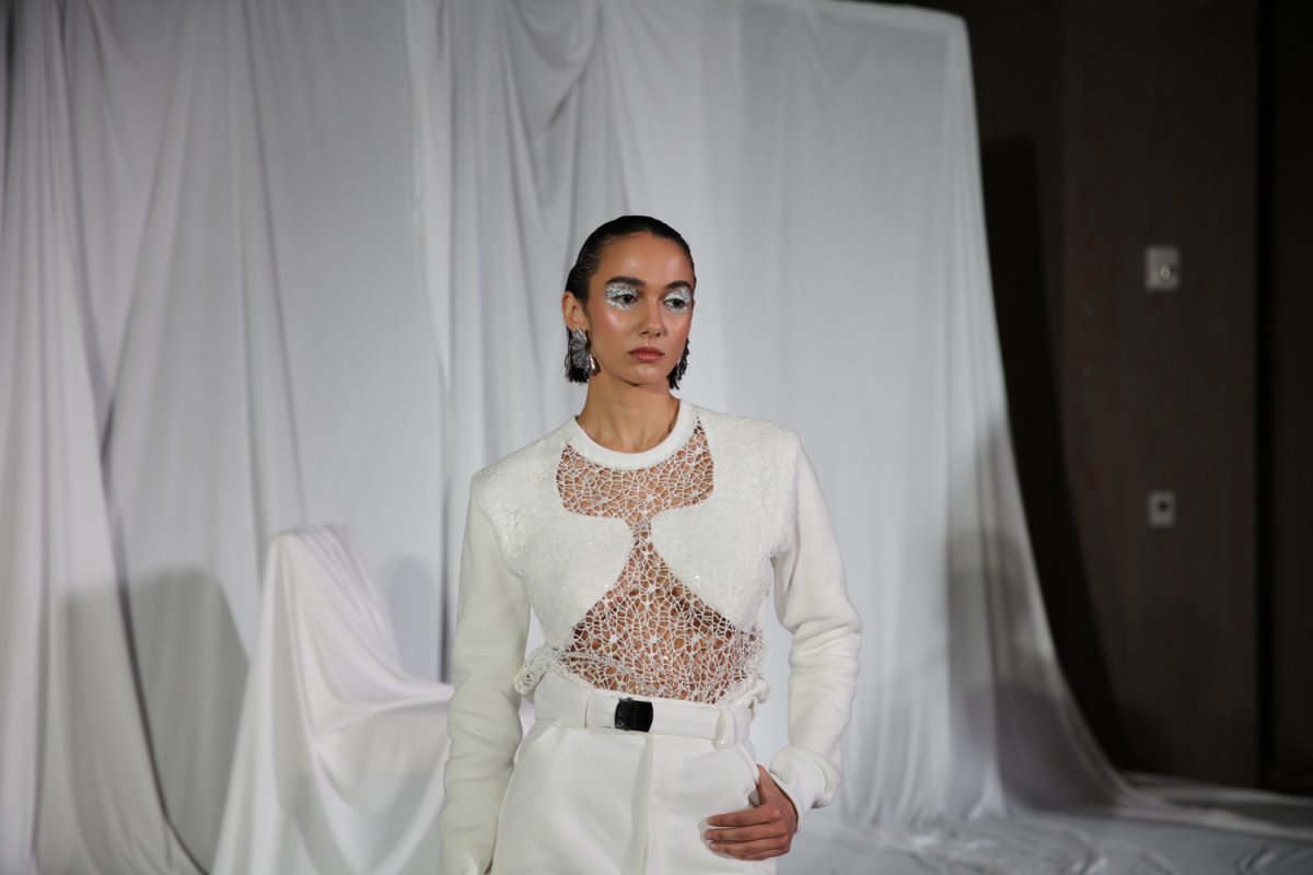 A model wears a white glittery top with a geometric cutout and white pants.