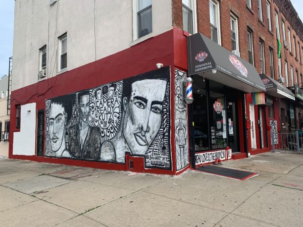 Side view of the exterior of a hair salon that has a black and white mural of three people on a red wall. The mural says “CAMERA READY KUTZ INC 2007.”