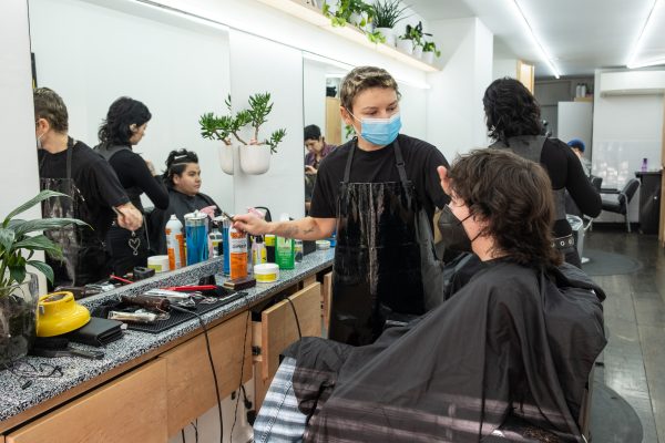 A person in a blue face mask and black apron is styling a client’s hair who is wearing a black barber’s cape and a black face mask. Hair equipment lines the tabletop in front of them.