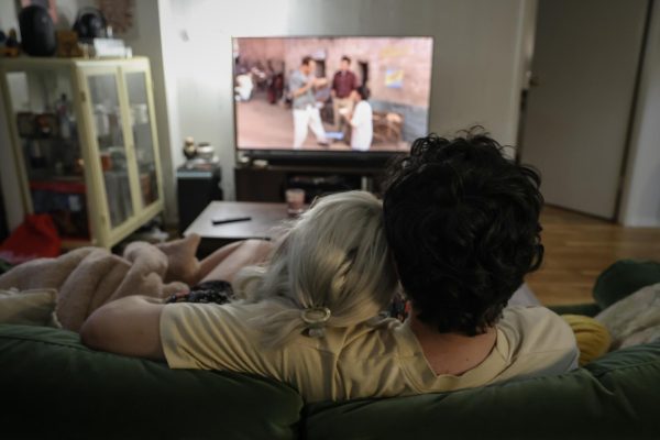 A woman with white hair rests her head on a man's shoulder. They are sitting on a couch watching television.