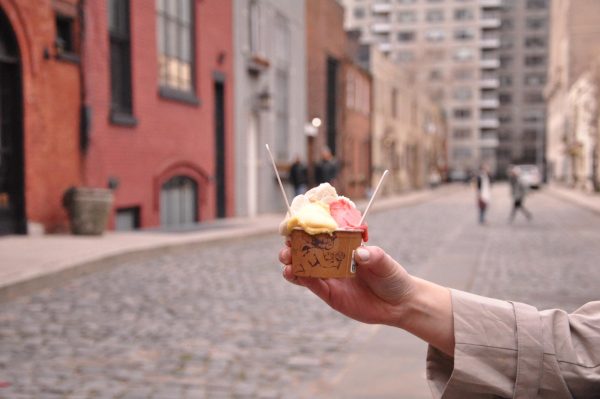 A hand holds yellow, pink and white gelato with two spoons sticking out in front of colorful buildings.