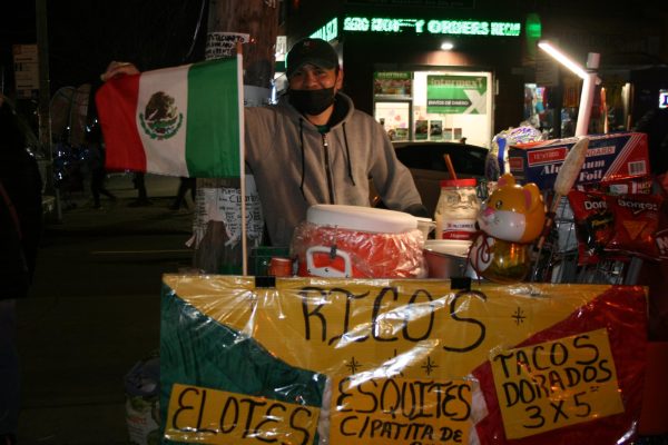 A food stand owner holds a Mexican flag on his cart. The cart is labeled with a sign advertising tacos and elote.