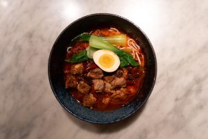 A dark blue bowl filled with red broth, pork ribs, vegetables, rice noodles and half an egg placed on a white marble table.