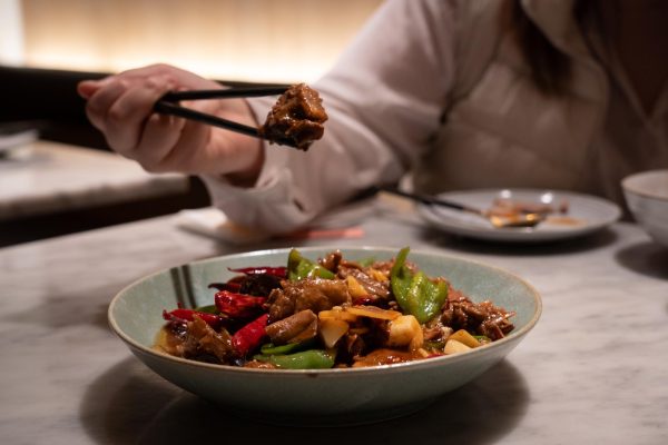 A person picking up a piece of duck with a pair of chopsticks from a dish that is filled with duck and red and green peppers.