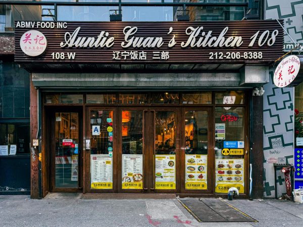 The exterior of Auntie Guan’s Kitchen, which has a brown exterior with white lettering and large windows. In the windows, are large printouts of the menu with pictures.
