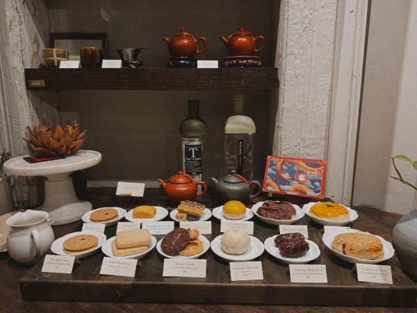 Twelve plates of pastries are set out, in front of an illustration of a dragon, two teapots and two glass bottles. On the shelf are two dark red teapots, two brown tea cups and a filter.