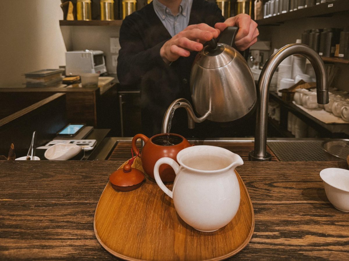 A man pours hot water from a metallic kettle into a teapot on a wooden tray. Beside the teapot is a white, clay pitcher.