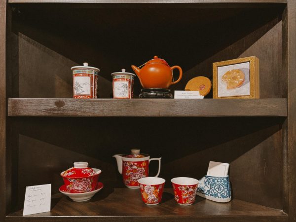 Two shelves display tea sets. The top shelf has a dark red teapot, two red cups and a box of pastries. The bottom shelf has two red teapots, two small red cups with floral patterns and a blue and white ceramic shoe.