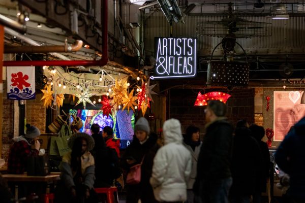 The interior of a marketplace decorated with hanging lights. A lit neon sign that says “ARTISTS & FLEAS” is hung from the ceiling, while a crowd of people are sitting by tables or walking by.