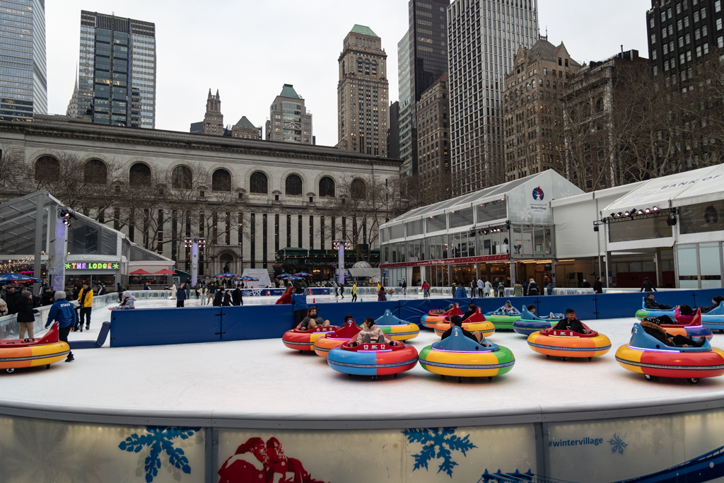 Many people are ice skating near the rink. The rink is decorated with snowflakes and lights. Half of the rink is full of colorful bumper cars.