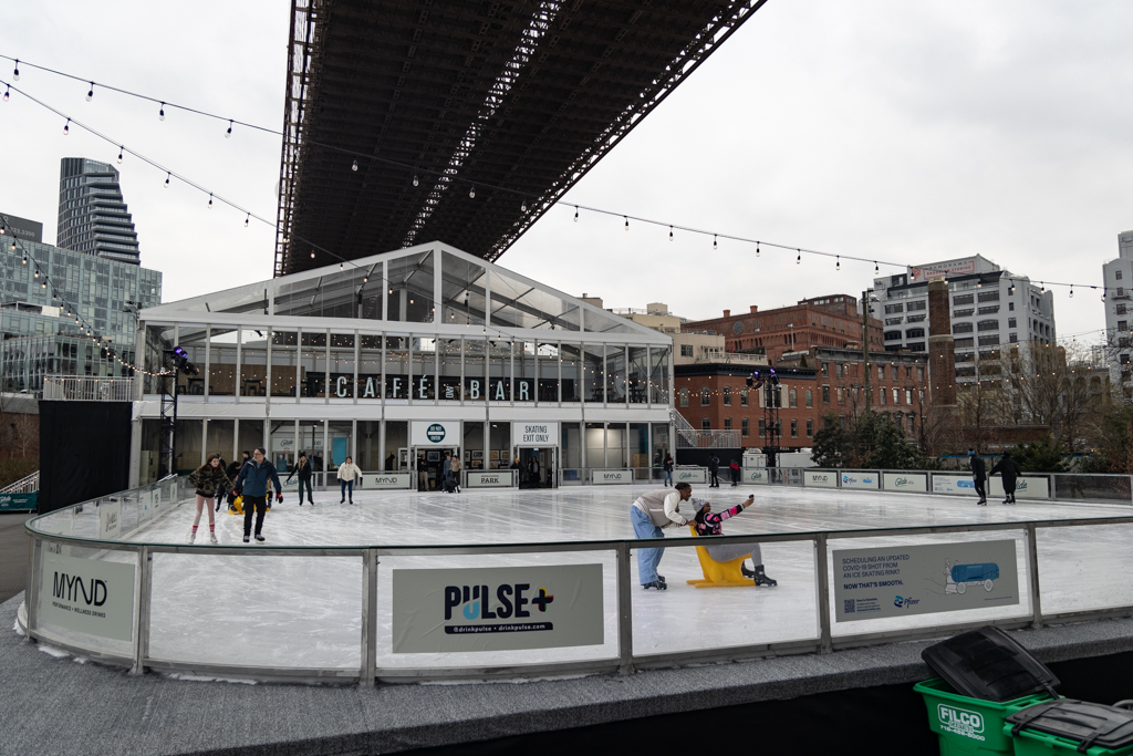 Several people skating on a medium-sized rink under a bridge. The rink is decorated with string lights and the names of sponsoring brands are displayed on the sides of the rink. Behind the rink is a building labeled “Cafe and Bar.”