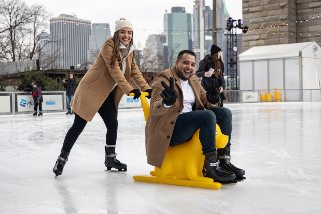 Two people are laughing while skating on the rink. One is sitting on a bright yellow glider shaped like a dog that is meant to help kids learn how to skate. That person is holding up two peace signs while the other pushes them.