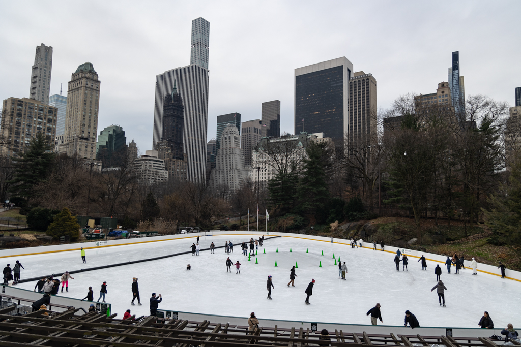 A wide shot of the ice-skating rink and the surrounding cityscape. Many people skate around the rink in a circle while others on the side film them.