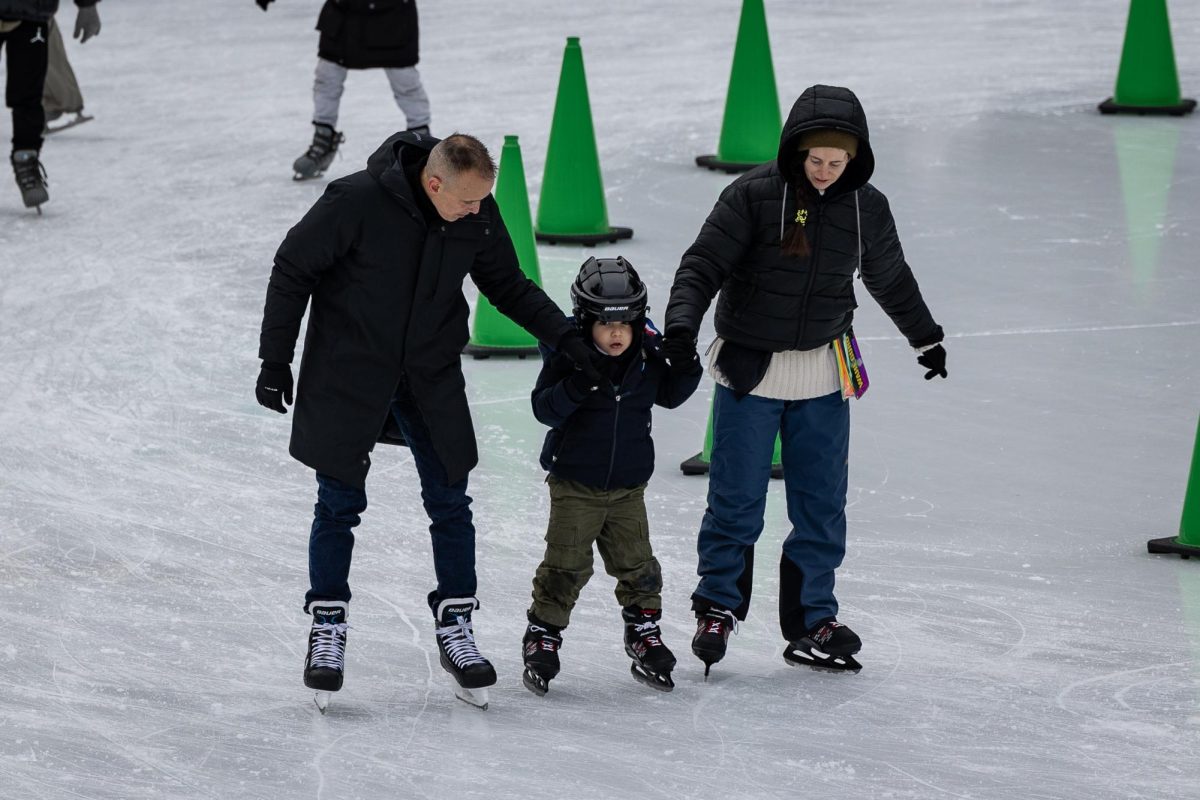 Two parents hold each hand of their young child as they ice skate around the rink. Each of them are bundled up with coats and the child has a helmet.