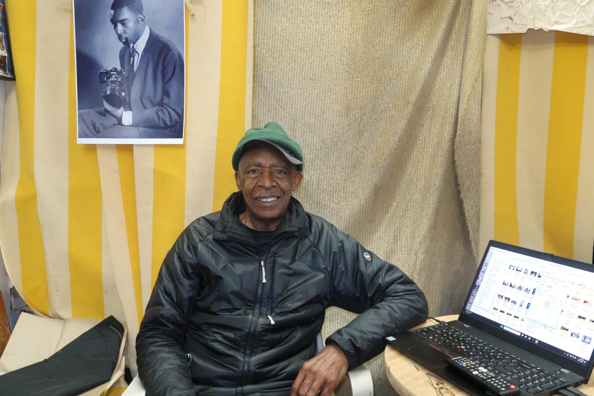 Image of a black man sitting in front of a yellow and white striped wall and next to an open computer.