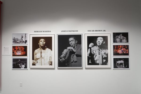 A white wall displaying photographs of musicians Miriam Makeba, John Coltrane, and Oscar Brown Jr. performing. “MIRIAM MAKEBA,” “JOHN COLTRANE,” “OSCAR BROWN JR” surrounded by smaller photographs of black musicians.