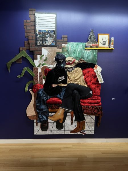 An installation placed on a dark blue wall, where a figure wearing a black shirt, a pair of blue jeans and a black cap and a person wearing a yellow shirt, a pair of black trousers and a beige hat hold each other while they sit on a red couch in an apartment.