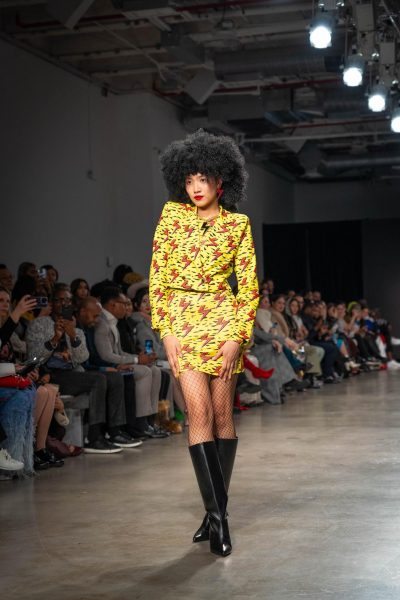 A model walking down a runway with a matching yellow and black set decorated with red lightning bolts and a large gold and black lightning chain necklace.