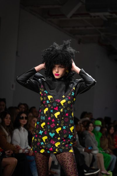 A model walking down a runway wearing a black leather dress with red, yellow and blue patterns and a pair of pink and black stockings places her hands on the back of her head.