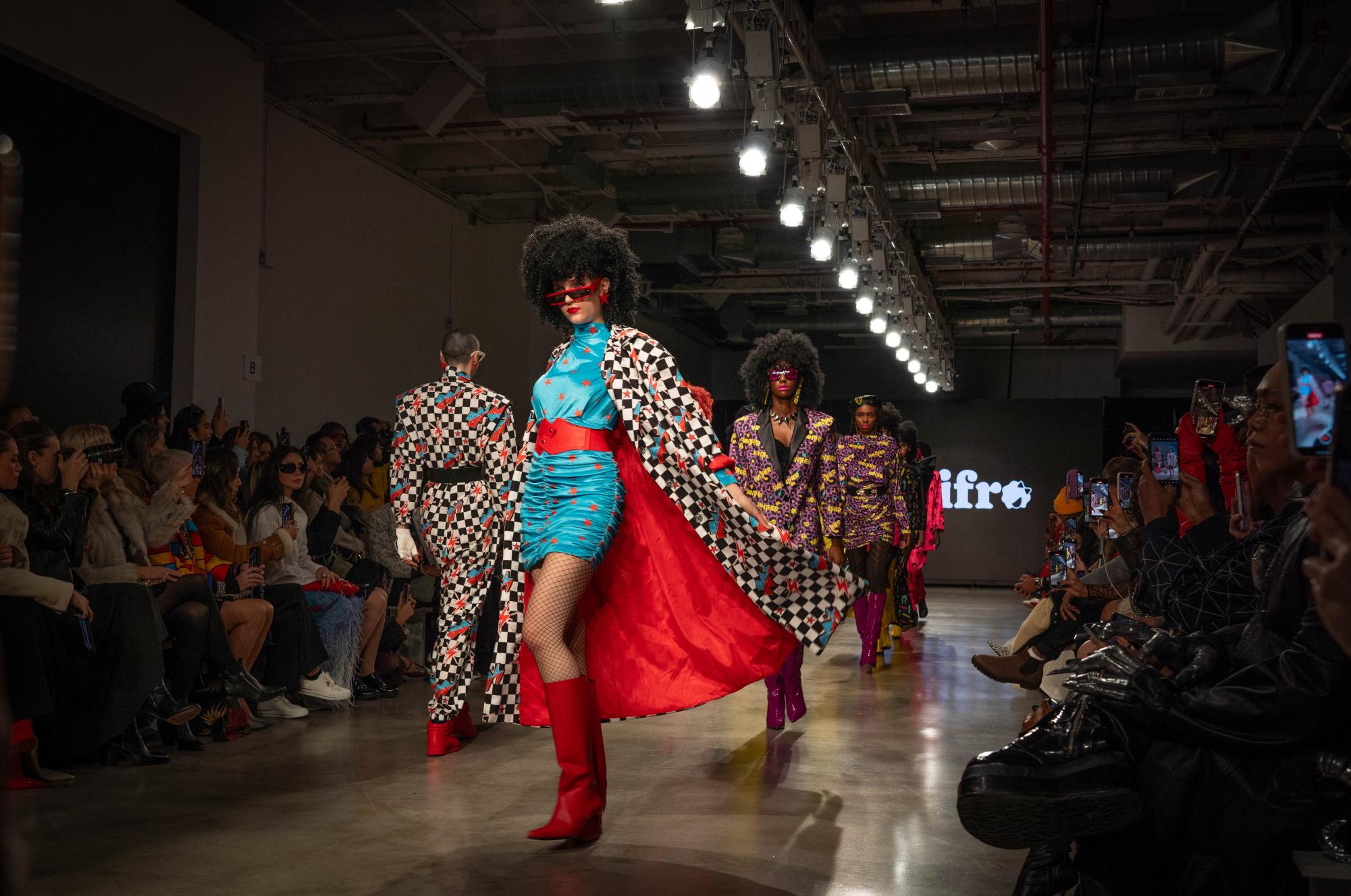A group of models walks down a runway in a line. At the front of the runway, a model opens a black and white checkered cape to reveal a blue ruched mini dress with red stars.