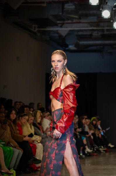 A model walking down a runway in a matching black-and-red patterned fabric set. The top has red leather sleeves and the bottom is a maxi skirt made of patterned fabric.