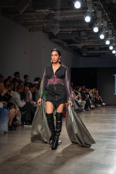 A model walking down a runway in a black dress with sleeves that flow to the ground, thigh-high black boots and a corset.