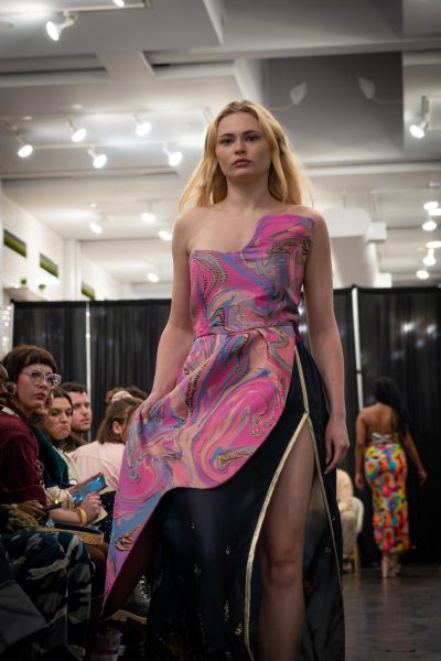 A model walks down a runway wearing a pink, orange, blue and gold sleeveless dress with a black and gold slit.