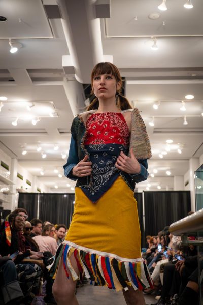 A model poses with a bandana halter top and blue woven jacket. They wear a yellow skirt with colorful tassels at the bottom.