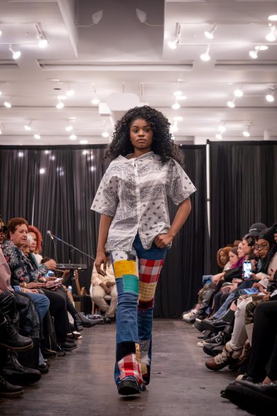 A model walks down a runway with a white paisley pattern shirt and patchwork jeans.