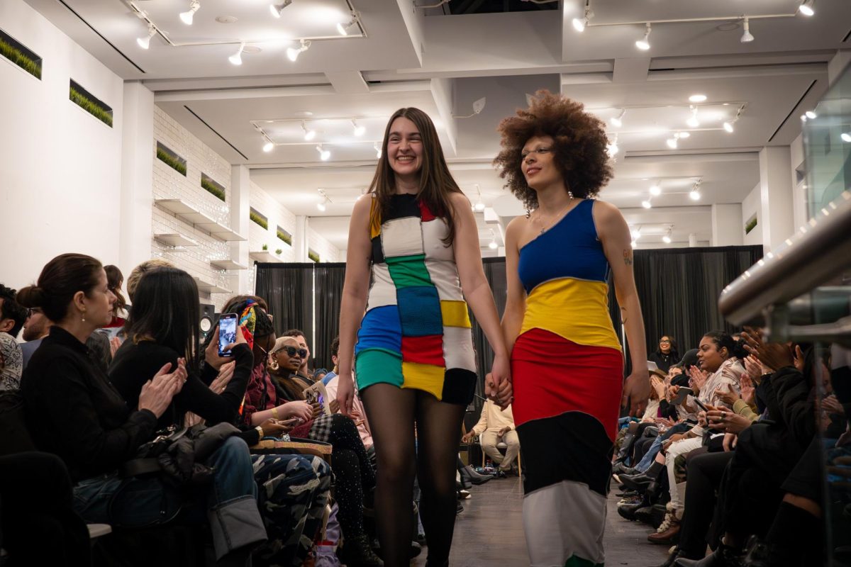 Two people walk down the runway. One wears a quilt patchwork dress and the other wears a striped colorblocked dress.
