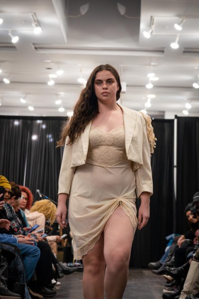 A model walks down the runway wearing a beige dress and a cropped beige blazer.