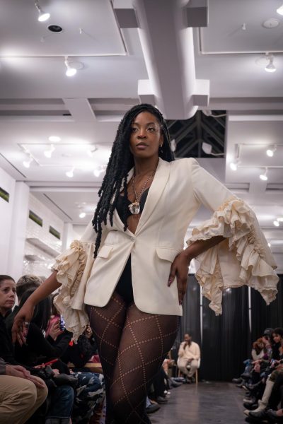 A model poses on a runway wearing a white jacket with ruffled sleeves, a pair of black lace stockings and a glass and metal necklace.