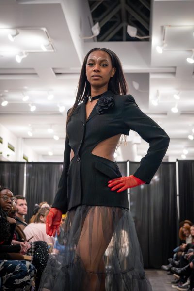 A model poses on a runway in a black blazer-like dress with cutouts with a sheer skirt coming off the bottom.
