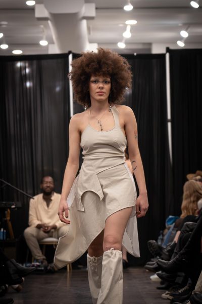 A model walks down a runway in a beige dress with uneven layers and beige fabric boots.