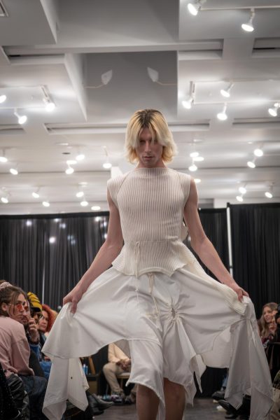 A model walks down a runway wearing a white sheer top with stripes and a white skirt with uneven layers.