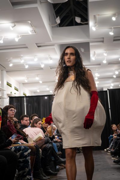 A model walks down a runway in a bulbous beige dress with an orange floral pattern at the top and red gloves.
